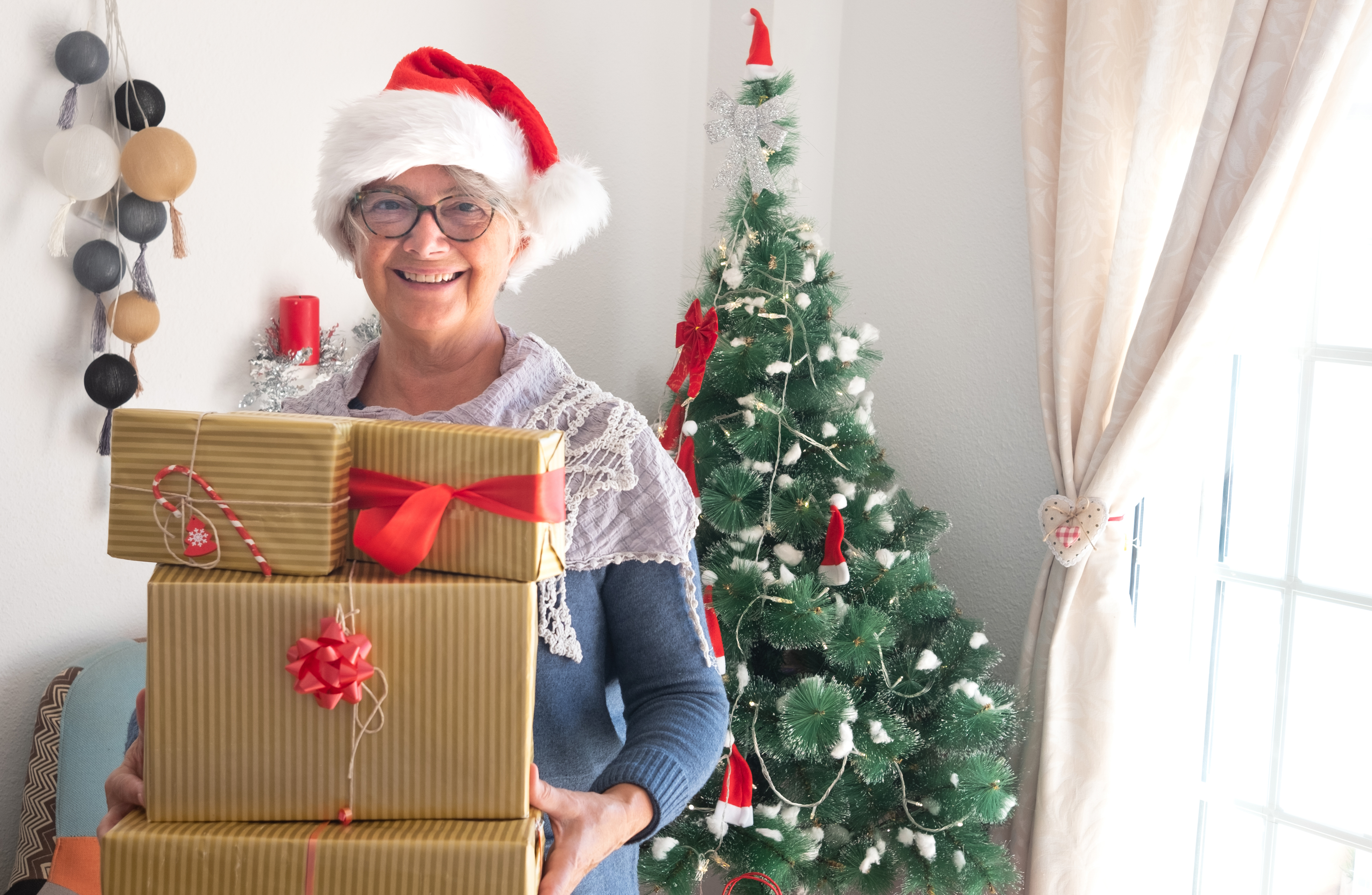 Happy people holding Christmas presents. Christmas tree on background. Senior female with Santa's cap and smiles